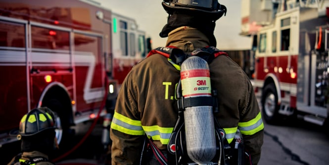 Un pompier, vu de dos, portant un équipement de protection complet, y compris un casque et un réservoir d’air Scott 3M sur le dos.
