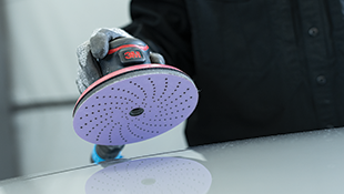 Une main gantée manie une ponceuse orbitale 3M munie d’un disque de ponçage perforé contre une surface lisse.
