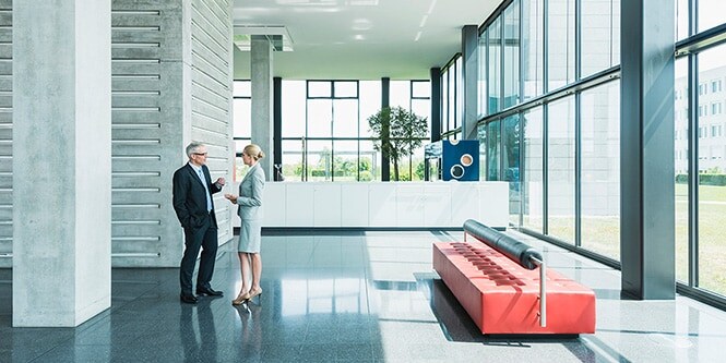 Vue panoramique du hall d’entrée d’un immeuble de bureaux contemporain, bordé de parois vitrées où deux hommes d’affaires échangent.

