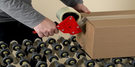 Worker uses a handheld red tape dispenser to seal a cardboard box with clear packing tape while it rests on a roller conveyor.
