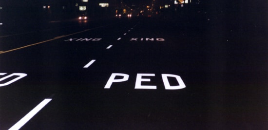 White illuminated pavement markings on dark road at night, reading 'PED XING'.
