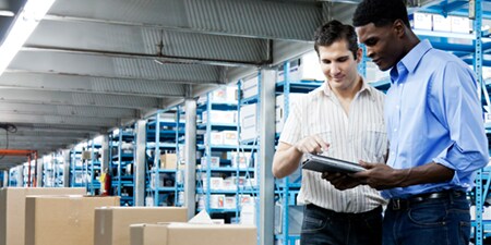 Deux employés d’entrepôt examinent une tablette dans une allée de stockage, tandis que de grands rayonnages métalliques remplis de boîtes étiquetées s’étendent en arrière-plan et que des cartons scellés sont empilés au premier plan.
