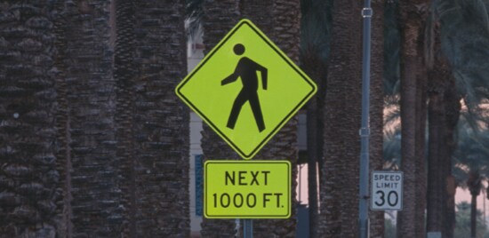 Standalone sign with yellow background and black image of two people crossing street and words 'school crossing'.

