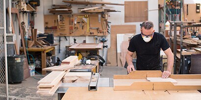 Un artisan à l’œuvre dans un atelier de menuiserie, équipé d’un masque de protection.
